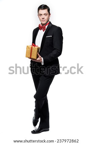 New year's eve fashion man wearing black dinner jacket with big red bow tie. Holding gold present. Isolated against white.