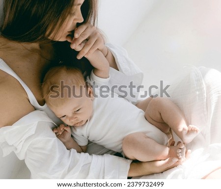 Young woman with baby in her arms. Blonde girl in white clothes smiling to your child on white background. Happy motherhood and breastfeeding concept. Photos with sun glare, soft focus, overexposure.