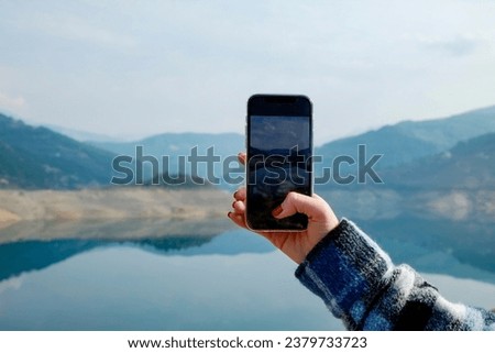 Cropped shot of woman's hand taking pictures of beautiful view. Mountain lake on a foggy cloudy day. Close up, copy space, background.