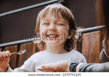 A beautiful little boy, a happy child, eats rice porridge with a spoon in his hand with the help of his mother. Photography, close-up portrait, baby food, feeding.