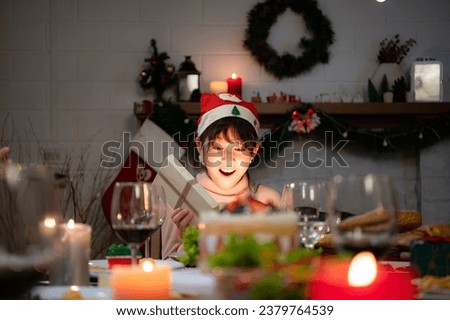 Cute little girl in Santa hat opening Christmas present and smiling.