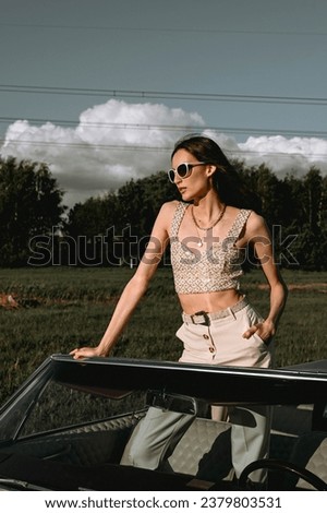 A beautiful woman stands in a convertible in a field. Dressed in white pants, a white top, and black sunglasses. Beautiful and stylish look.