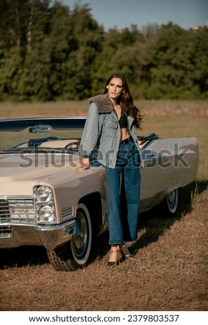 A beautiful woman stands next to a convertible in a field. Dressed in jeans and a jacket with fur. Beautiful and stylish look.