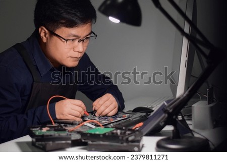 Young man wearing glasses who is a computer technician A laptop motherboard repairman uses an IC meter to find defects on the motherboard to repair on his work table and has a lamp for illumination.