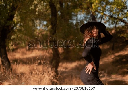 Happy gothic young woman in a Halloween witch costume with a hat standing and smiling against the background of an autumn forest, a party in the forest, a party outfit for the Halloween holiday