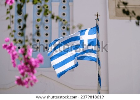 View of the greek flag waving in the air next to a bougainvillea and a whitewashed church in the background in Ios Greece