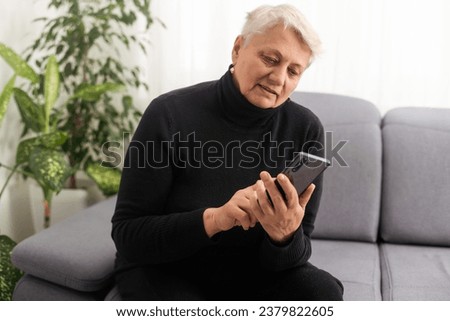 Relaxed mature old 60s woman, older middle aged female customer holding smartphone using mobile app, texting message, search ecommerce offers on cell phone technology device sitting on couch at home