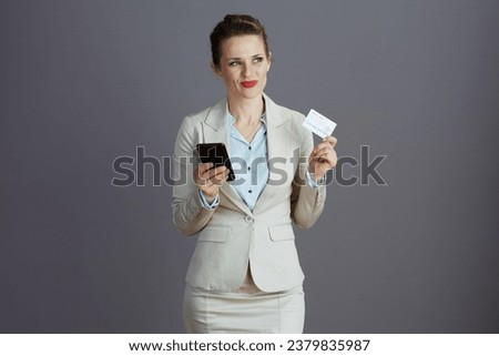 pensive middle aged small business owner woman in a light business suit with smartphone and credit card isolated on grey background.