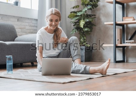 Sporty mature Caucasian woman athlete yogi trainer doing home workout, stretching, yoga classes online using laptop sitting on the floor at home Royalty-Free Stock Photo #2379853017