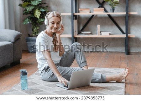 Happy mature Caucasian woman athlete yogi stretching her legs at home using laptop online tutorial workout, listening to the music after training cardio Royalty-Free Stock Photo #2379853775