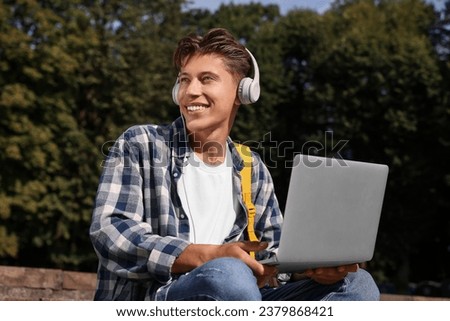 Happy young student with headphones studying with laptop on steps in park
