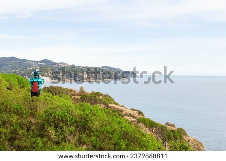 An unrecognizable adult person, dressed in winter clothes, is taking photo of seascape. He wears a red backpack, a green sweater and a blue wool hat. Lloret de Mar. Spain.