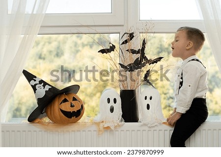 Halloween. Concept. A cute and cheerful boy sits near the window against the background of white ghosts and a vase with dry branches and black paper bats. Close-up.