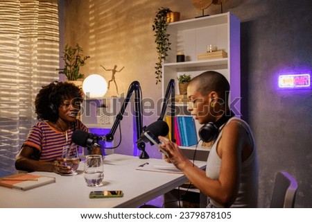 A happy multiracial woman is listening to the host who is interviewing her during the live stream in a podcast home studio. Multiracial gen z girls making a podcast at home while talking at the mic.
