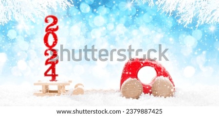 Red Wooden Car Pulling Sled with Wooden 2024 Numbers  in perspective on bright blue bokeh winter background with white snowy branches and falling snow.
