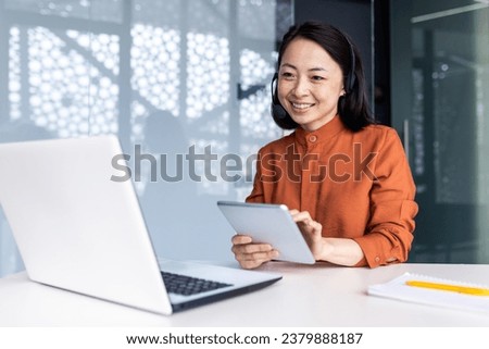 Young successful asian woman inside office at workplace with tablet computer and headset phone, woman advises buyers consumers remotely, business woman use laptop for video call. Royalty-Free Stock Photo #2379888187