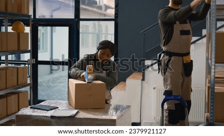 Dolly out shot of head of operations in warehouse supervising trainee sealing cardboard box parcels. Retail depository personnel properly securing goods to avoid damages during shipping Royalty-Free Stock Photo #2379921127