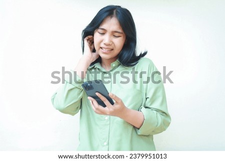 stressed anxious asian woman grabbing her neck confused using social media and online shopping on smartphone wearing green oversized clothes standing over isolated white background, feeling anxiety
