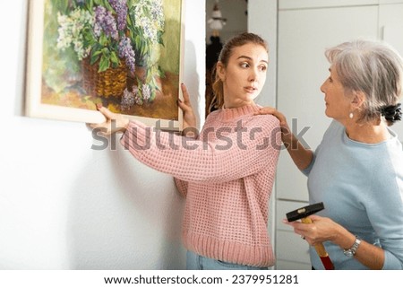 Interested senior woman with her adult daughter hanging framed picture on wall at home.