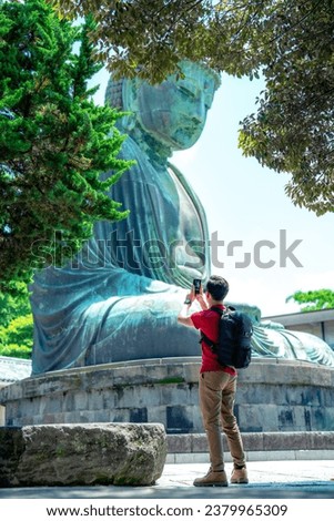 Full body side view of Hispanic unrecognizable young male in casual clothes with backpack while taking picture with smartphone of Kotoku-in Buddhist temple structure in Kamakura Tokyo, Japan