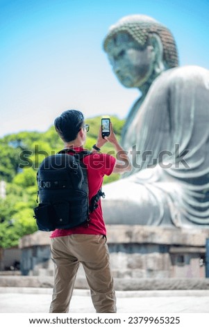 Full body view of Hispanic unrecognizable young male in casual clothes with backpack while taking picture with smartphone of Kotoku-in Buddhist temple structure in Kamakura Tokyo, Japan