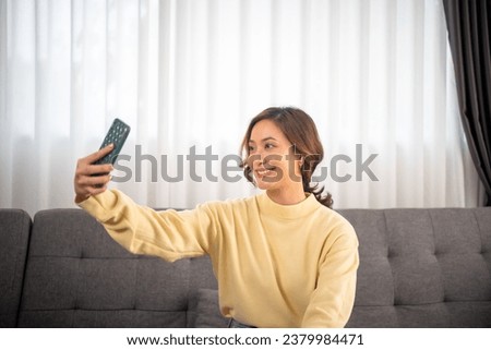 An Asian girl with a big smile sits in the living room. She has fun taking cheerful use smartphone video conference selfies and looking at the camera
