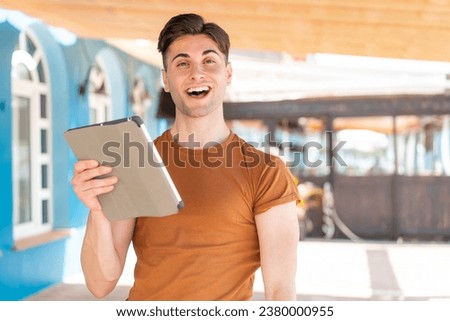 Young handsome man holding a tablet at outdoors with surprise and shocked facial expression