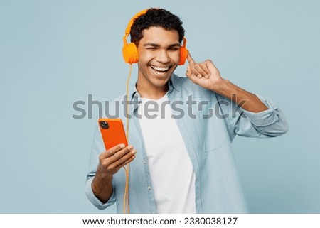 Young cheerful man of African American ethnicity he wears shirt casual clothes listen to music in headphones use mobile cell phone isolated on plain pastel light blue cyan background studio portrait Royalty-Free Stock Photo #2380038127