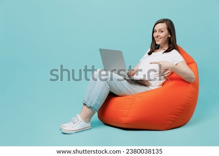 Full body young woman 20s she wear white t-shirt sit in bag chair hold use work point index finger on laptop pc computer isolated on plain pastel light blue cyan background. People lifestyle concept