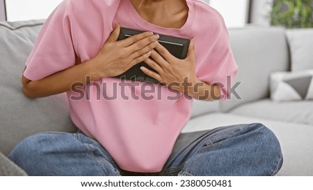 Relaxed hispanic woman comfortably sitting on living room sofa, holding cherished photo frame close to her chest