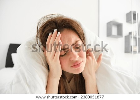 Portrait of woman with headache, lying in bed and touching head with frustrated, frowning face, does not feel well, stays in bedroom as having migraine in morning. Royalty-Free Stock Photo #2380051877