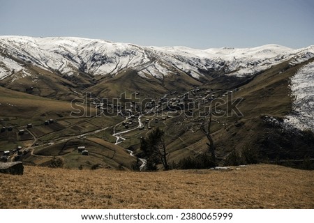 Rural sunny landscape: snowy mountains, meadows, a river, and a village view from above. High-quality photo for website design, postcards, banners, and travel product advertising