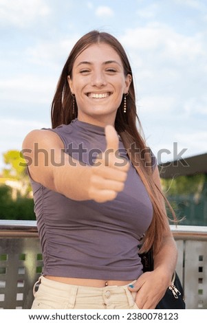 Vertical portrait Young Caucasian woman pointing thumb up looking smiling at camera outdoors.