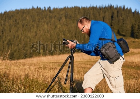 Professional photographer taking pictures in mountains. Man focusing his camera to take nice photo. Side view of young male adjusting camera on background of hills.