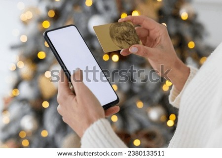 Close-up photo of young female hands using a phone with a white mock-up screen and holding a credit card against a background of a decorative Christmas tree during the New Year holidays.