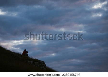 Nature photographer in the action. Man silhouette on mountain. Silhouette of a man photographer standing on top of mountain at sunrise cloudy background. The photographer with the tripod takes photos.