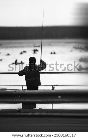 Silhouette of a teenager on a bridge with a fishing rod against the background of a water surface with boats with fishermen..