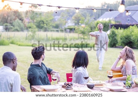 A young, attractive millennial man arrives at a garden dinner party or BBQ to find a multi-ethnic group of friends already enjoying wine and food around the table. As he approaches, the gathering Royalty-Free Stock Photo #2380160925