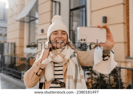 stylish young pretty woman walking in winter street wearing beige coat, knitted hat, scarf, smiling happy cold season fashion trend, taking selfie, making photo on phone