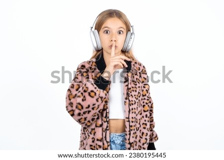caucasian kid girl wearing animal print jacket making hush gesture with finger on her lips wearing  wireless headphones. Be quiet. Royalty-Free Stock Photo #2380195445