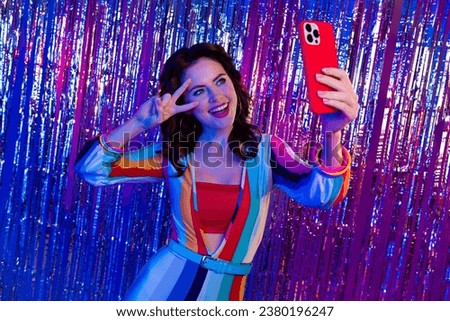 Portrait of charming funky person hold smart phone make selfie show v-sign backdrop tinsel decor isolated on multicolor neon background