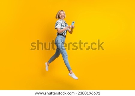 Full size photo of positive pretty gorgeous girl wear print blouse jeans flying holding smartphone isolated on yellow color background