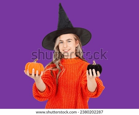 Halloween celebration concept. Portrait of smiling girl in black hat holding decorative pumpkins on violet background. Halloween decor.