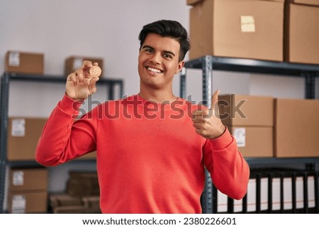 Hispanic man working at small business ecommerce holding bitcoin smiling happy and positive, thumb up doing excellent and approval sign 
