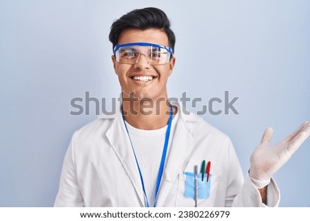 Hispanic man working as scientist smiling cheerful presenting and pointing with palm of hand looking at the camera. 