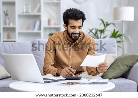 Young man working at home with documents and papers, homework, paying bills and loans, hispanic sitting on sofa in living room, using laptop and calculator. Royalty-Free Stock Photo #2380232647