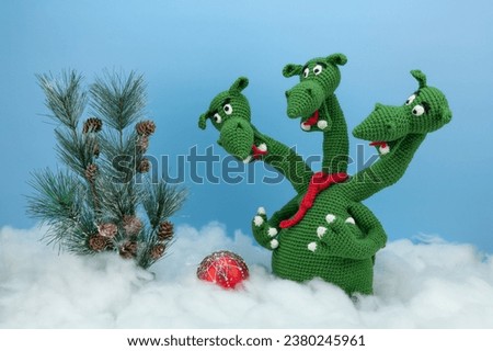 Three-headed dragon in the snow next to a Christmas tree and a red Christmas tree decoration. Knitted dragon.