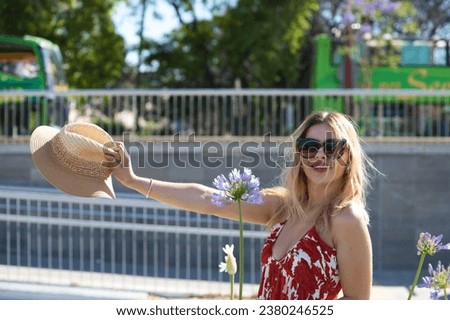 Young and beautiful blonde woman with sunglasses, raising her straw hat with her hand is among the violet flowers. The woman is on holiday and enjoys the city. The woman is happy