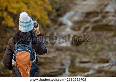 woman taking a photo of a waterfall in autumn with changing leaves on her mobile phone (hiker with backpack and jacket, hat takes cell phone picture of scenic nature and colorful foliage) from behind