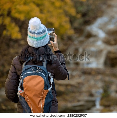 woman taking a photo of a waterfall in autumn with changing leaves on her mobile phone (hiker with backpack and jacket, hat takes cell phone picture of scenic nature and colorful foliage) from behind